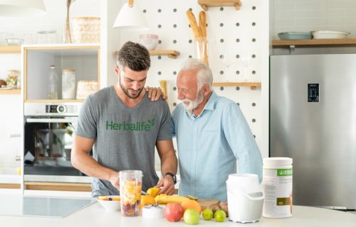Young man wearing Herbalife apparel preparing a protein shake with fruit for an older man in modern kitchen.This asset is being used as the hero image for the article, "How Much Protein Do You Need? Get the Right Intake and Protein Sources"
