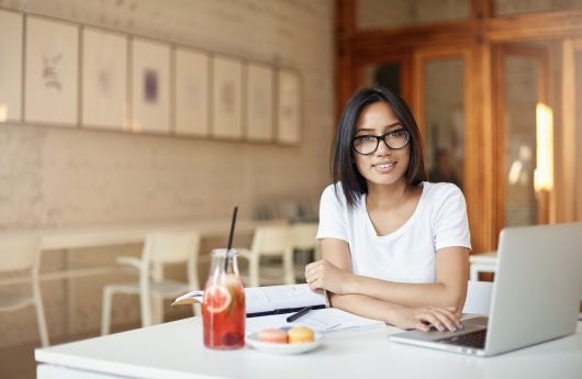 Young asian entrepreneur student working on laptop in library or open space cafe looking at camera